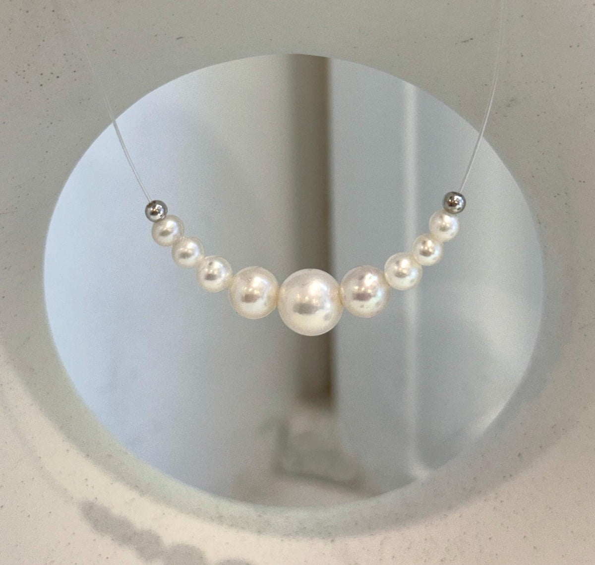 Nine white pearls arranged in a graduated curve on a clear nylon cord, displayed on a cream necklace stand.