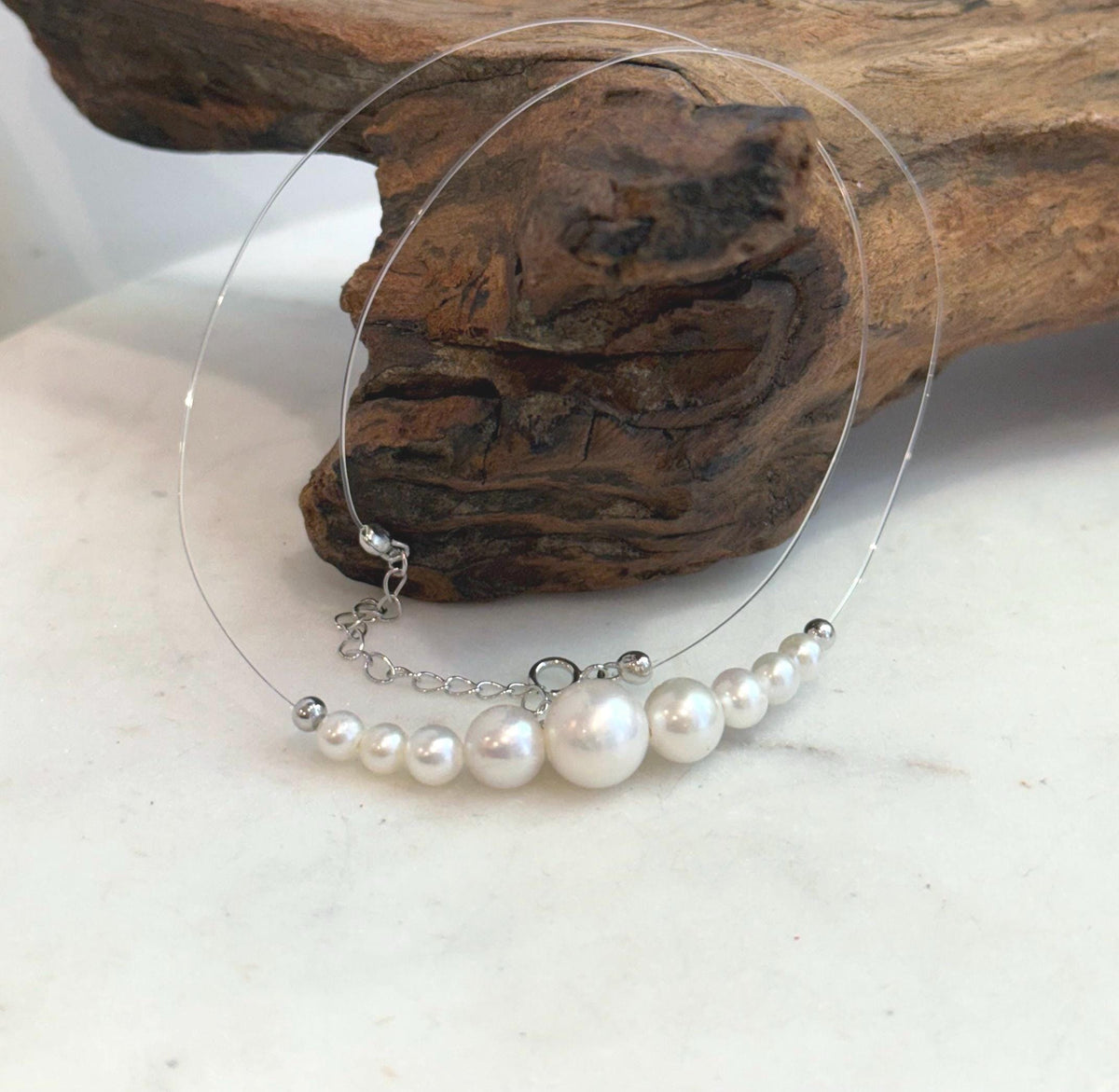 White pearl necklace laid flat on a pale surface, showing its clear nylon cord and extension chain.