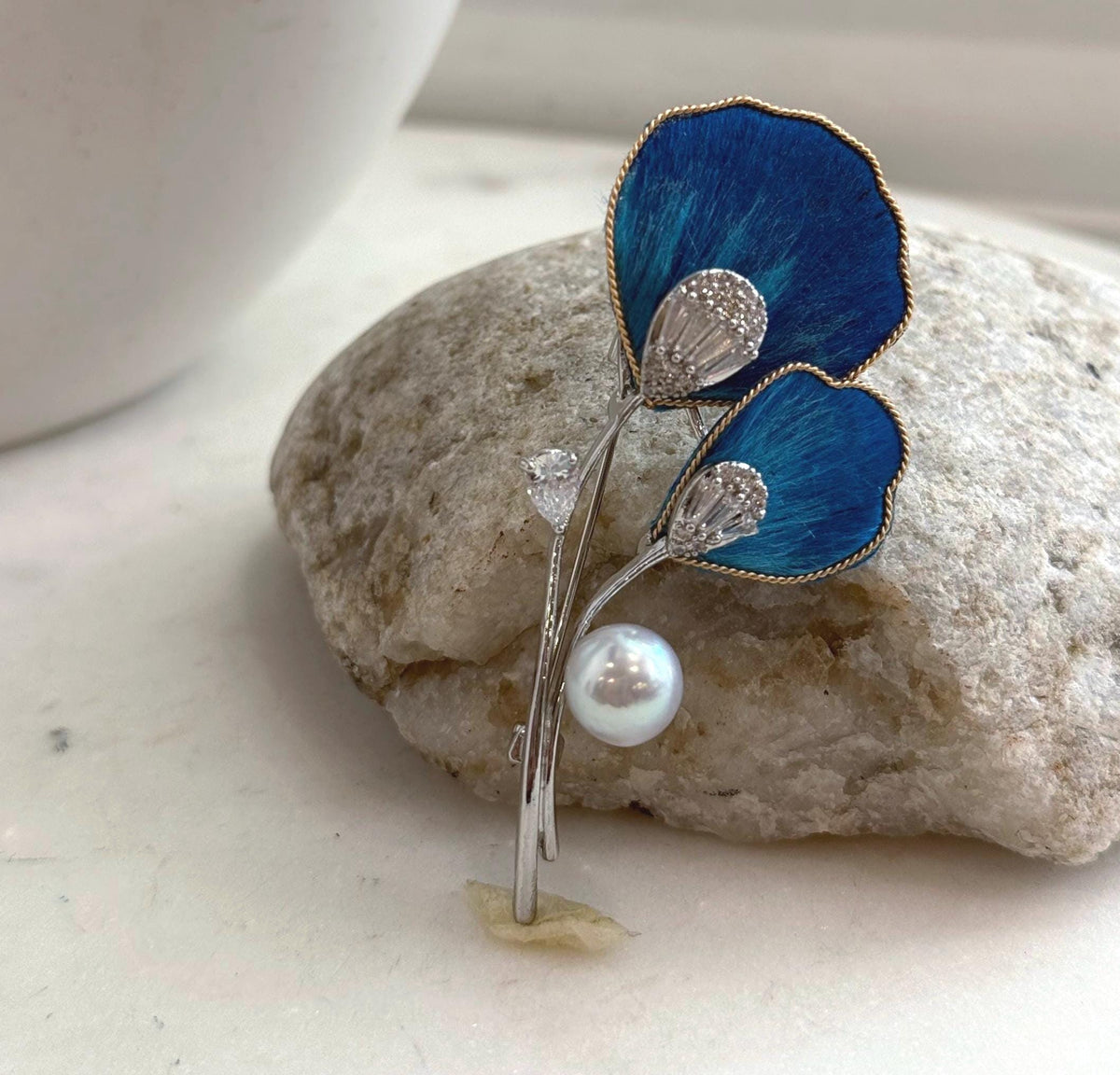 White Akoya pearl hanging beneath two vivid blue petals on a floral brooch pendant.