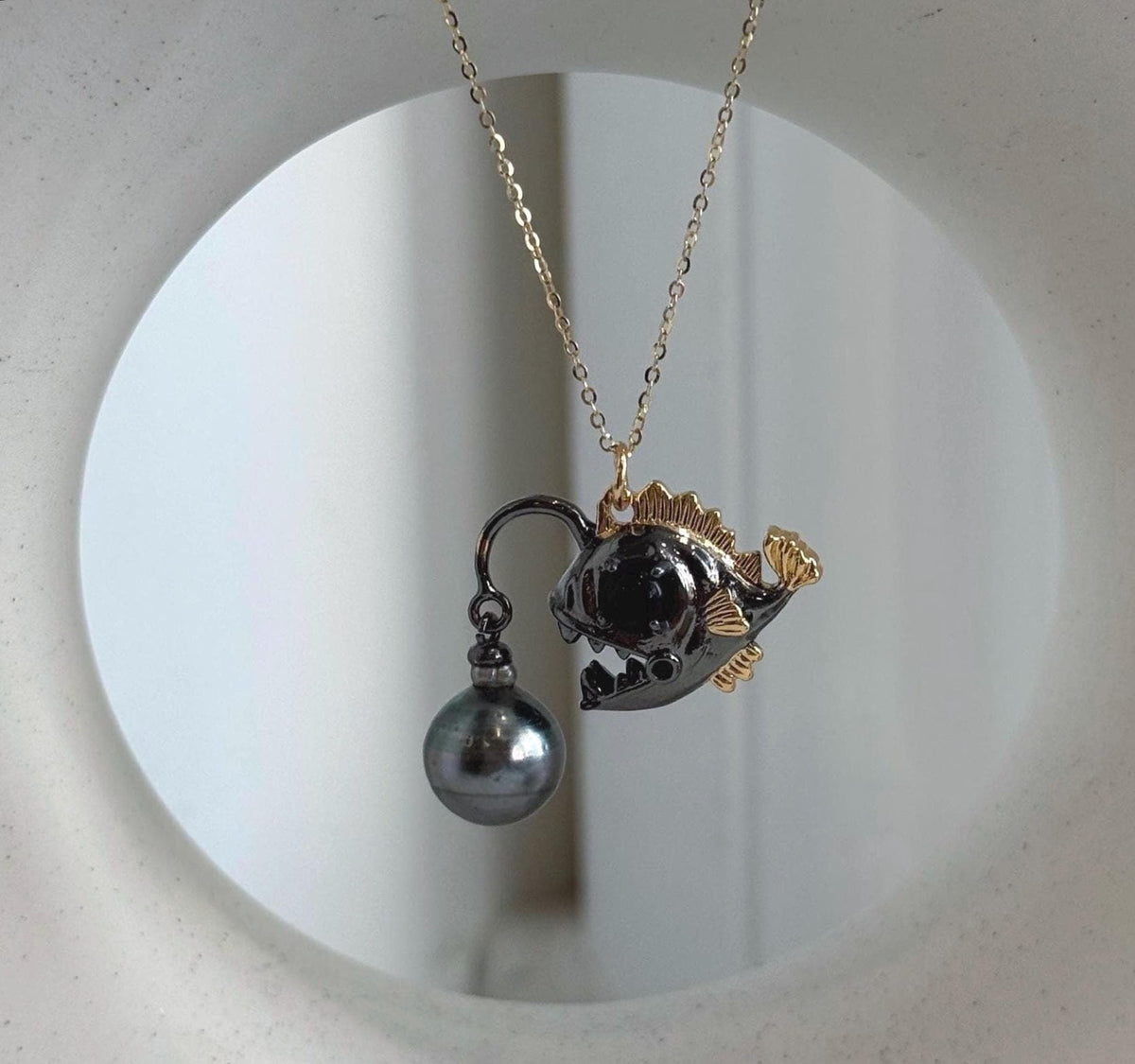 Dark metallic fish pendant with gold-tone fins and a dangling pearl.
