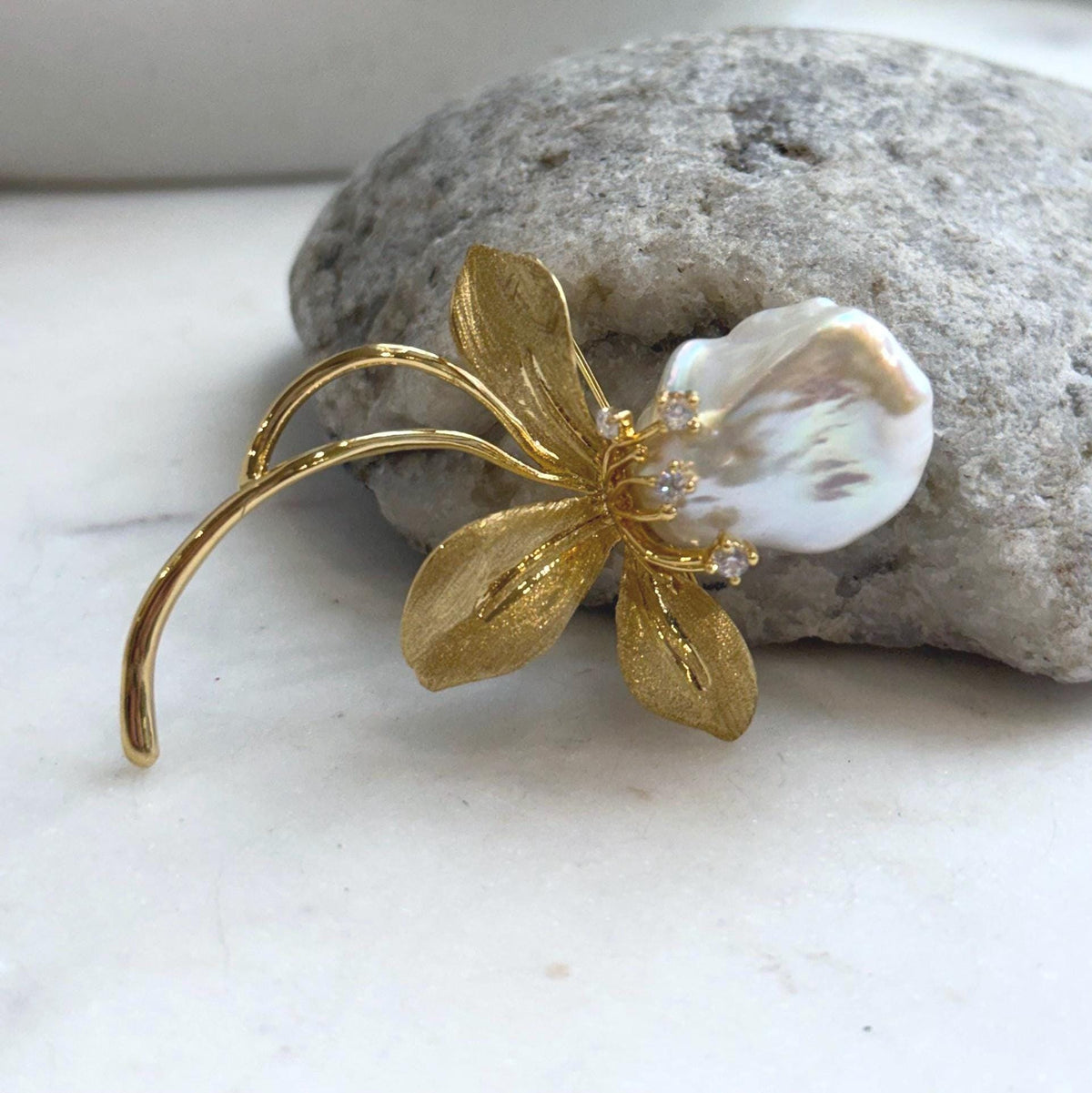 Iridescent baroque pearl above gold-tone flower petals and curved stems.