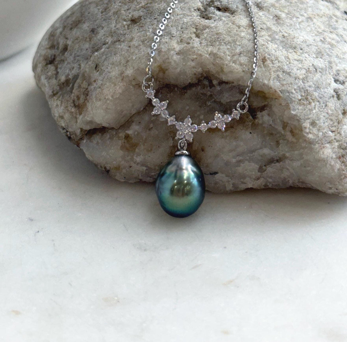 Tahitian pearl necklace displayed against a cream stand, showing its green pearl and sparkling floral centerpiece.