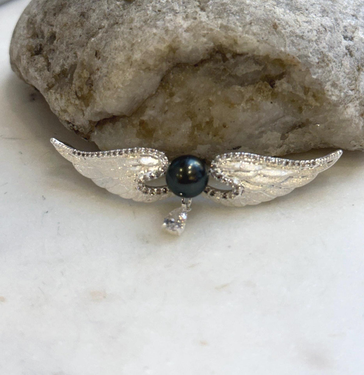 Black hunter green Tahitian pearl centered between silver-tone angel wings with a clear dangling accent.